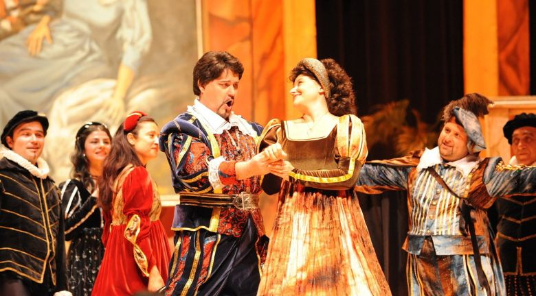 A theatrical performance featuring six actors in vibrant historical costumes, with two central figures singing and dancing together on stage.