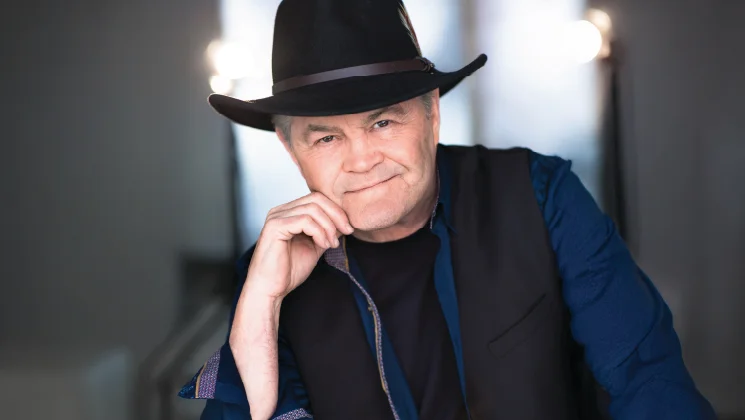 A smiling man wearing a black hat and a dark vest, leaning against a surface with a hand on his chin, set against a softly blurred background.
