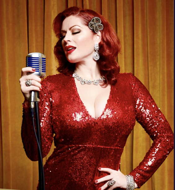 A glamorous woman with red hair wearing a sparkling red dress, holding a vintage-style microphone, against a golden curtain backdrop.