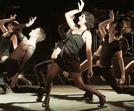 A group of dancers performing on stage, showcasing dynamic movements and dramatic poses. The lead dancer, dressed in a black dress, is in a striking pose while several others in black attire surround them.